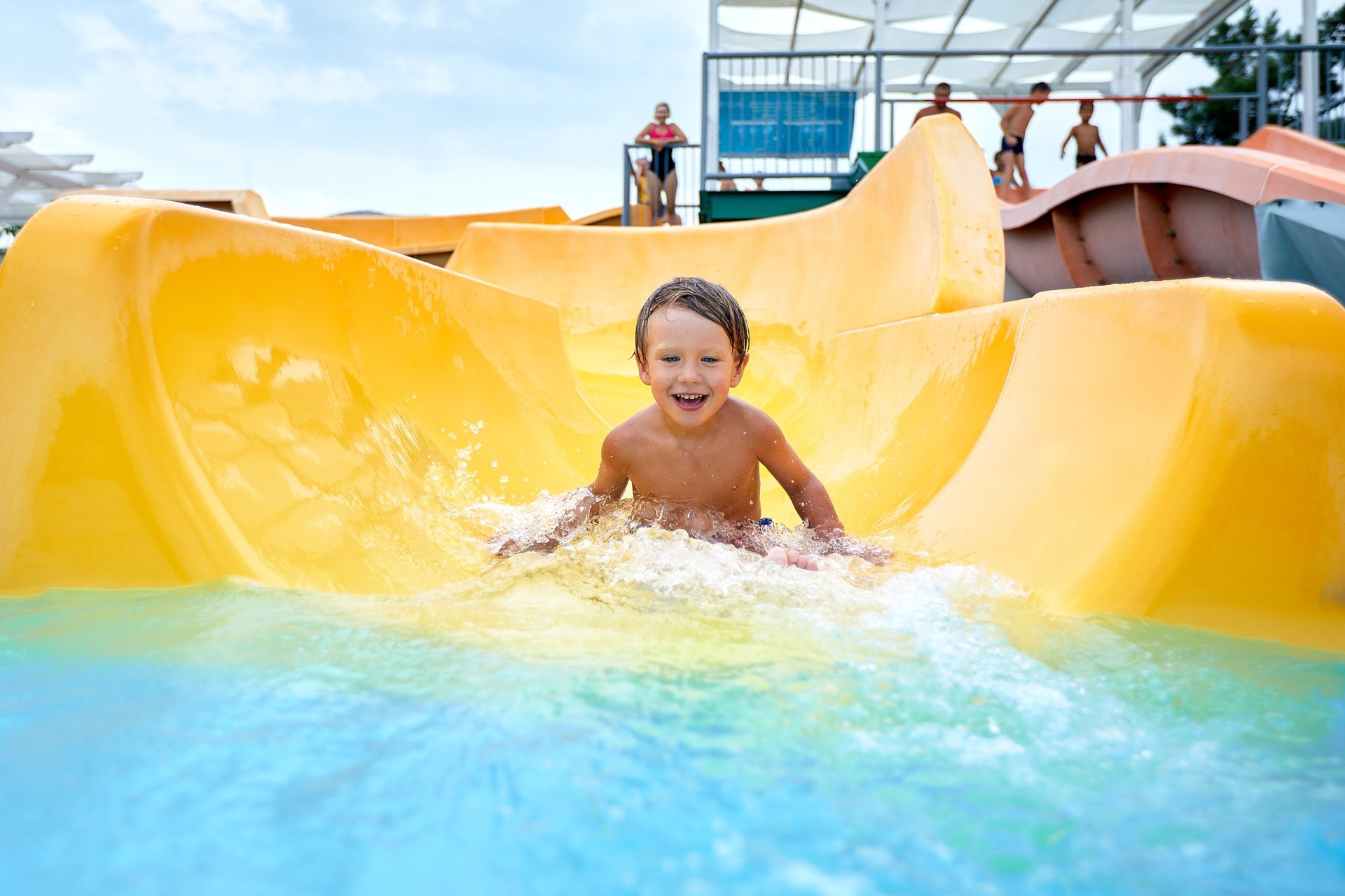 Cheerful blue-eyed kid on a water slide in the water park, a little boy merrily slides into the water along the slide in the amusement park.