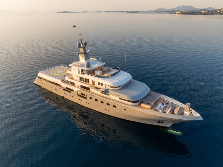 Aerial drone view of a luxury superyacht with a helipad, anchored in a calm turquoise bay at sunrise