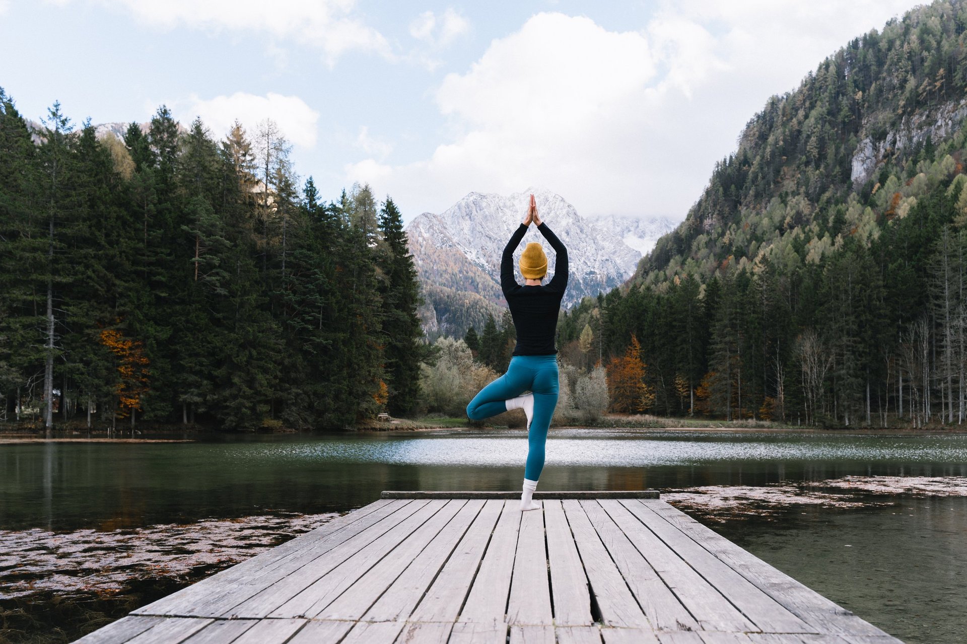 Mature fit woman practicing yoga on pier by lake overlooking green nature and mountains, concept of wellbeing and mindfulness.